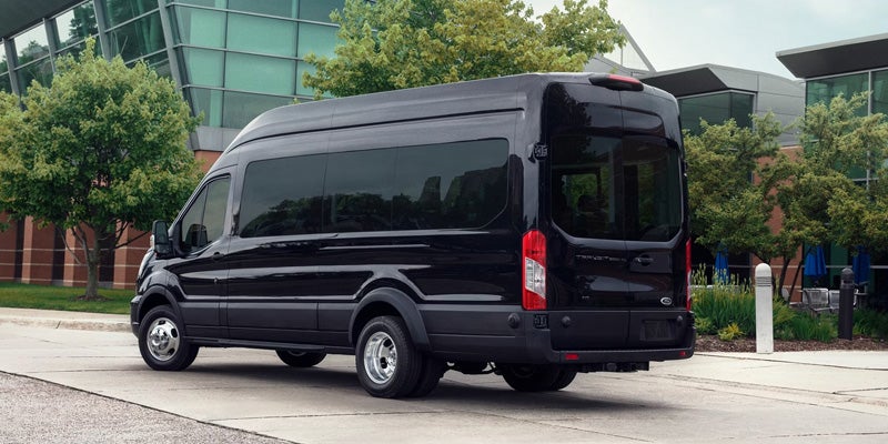 Rear-side view of a black Ford Transit cargo van parked on asphalt outside a modern building with green glass.